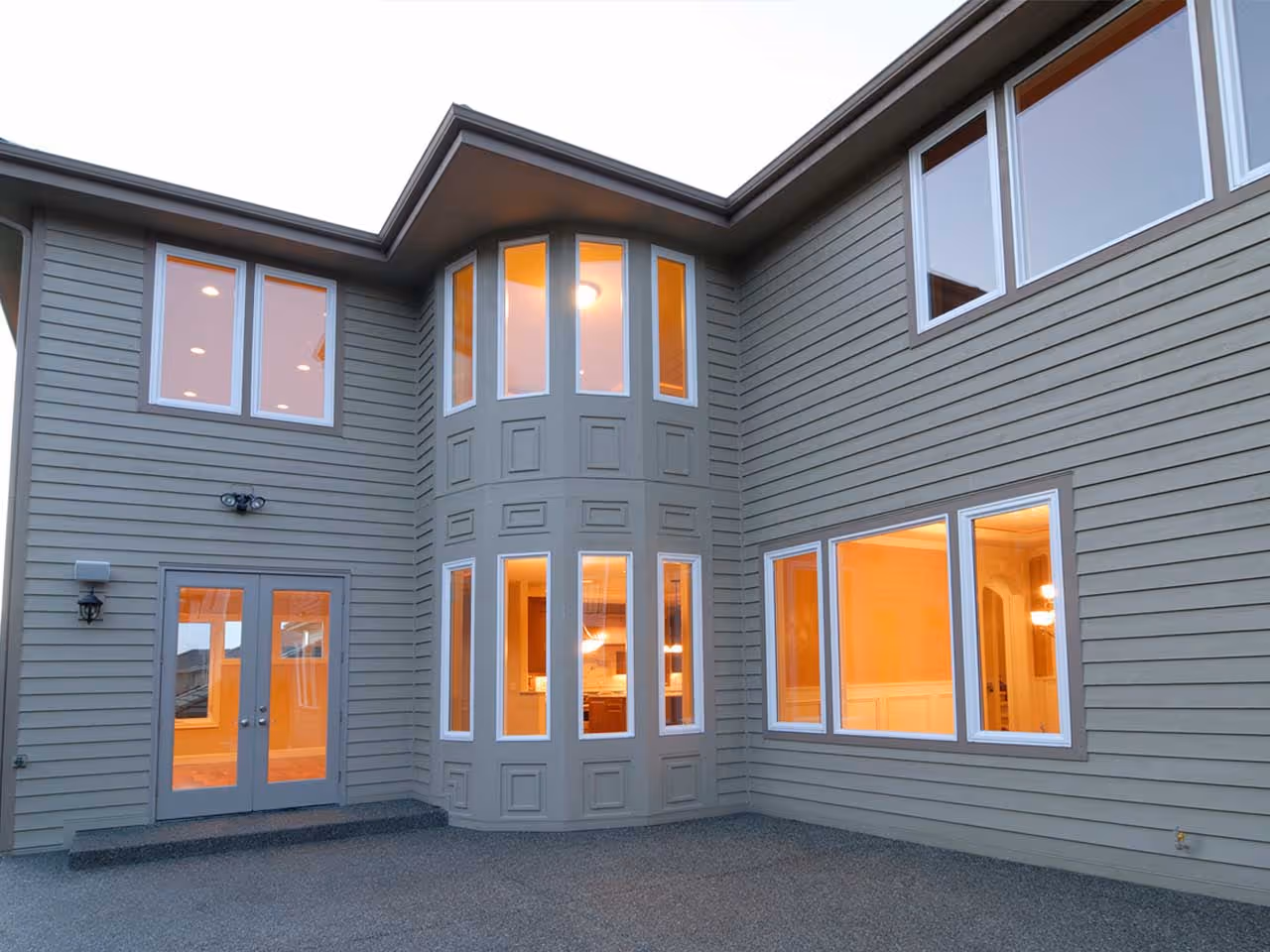 Exterior of a two-story gray house with lit windows and French doors opening to a concrete patio.