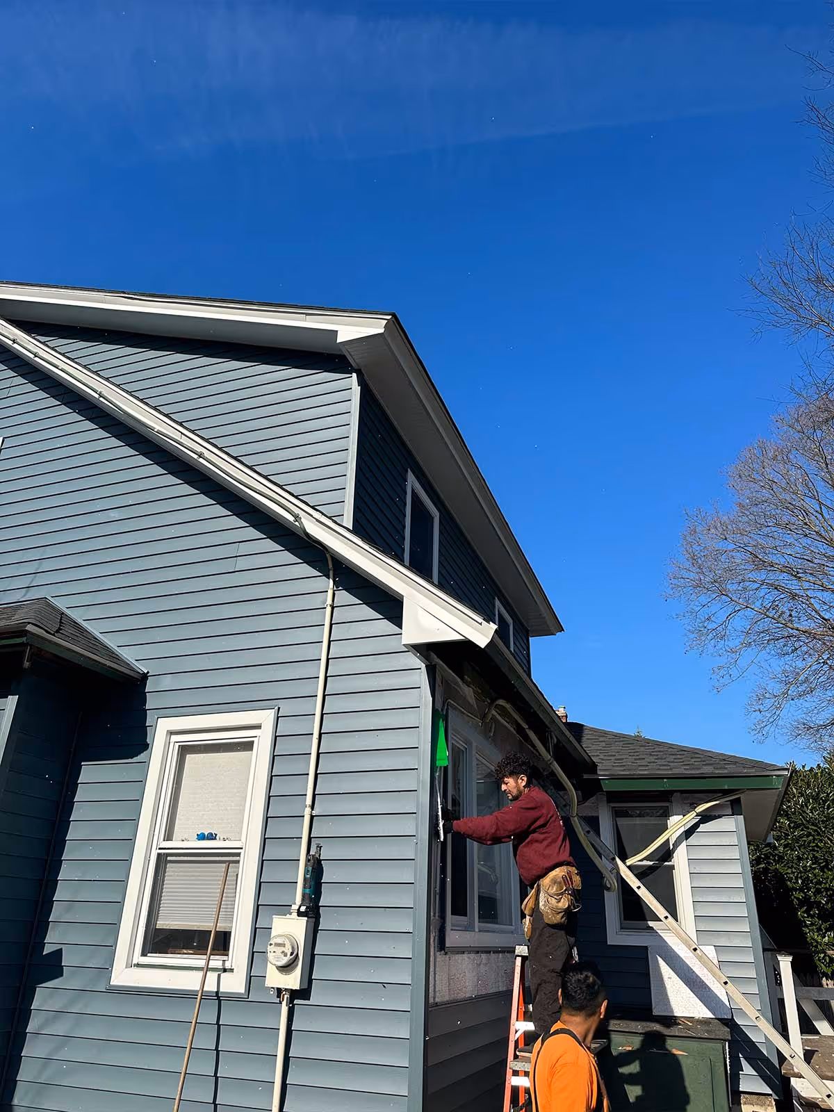Two workers installing or repairing a window on a blue vinyl-sided house under a clear blue sky.
