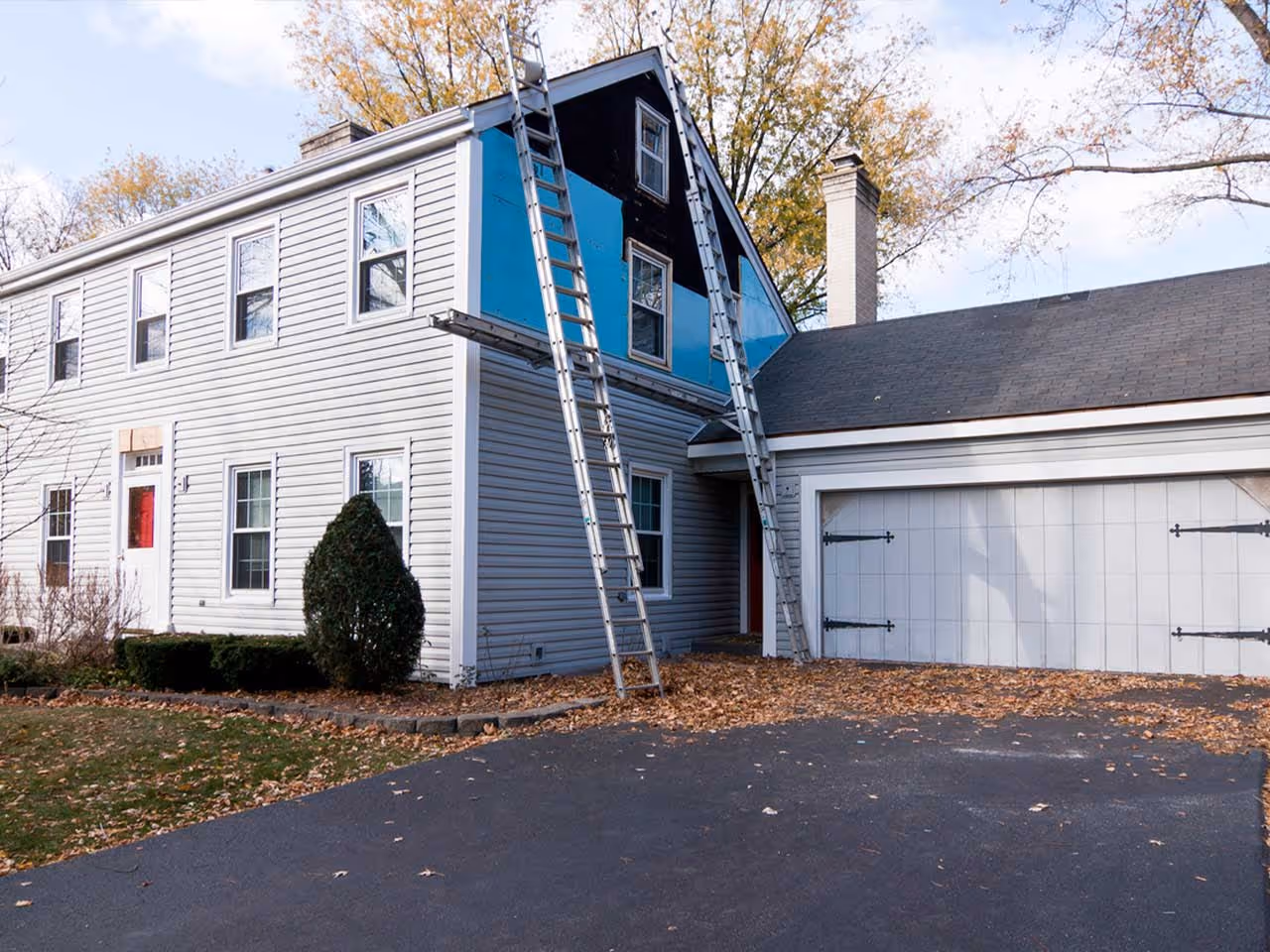 Exterior view of a two-story house with gray vinyl siding, two ladders leaning against the roof, and an attached garage.
