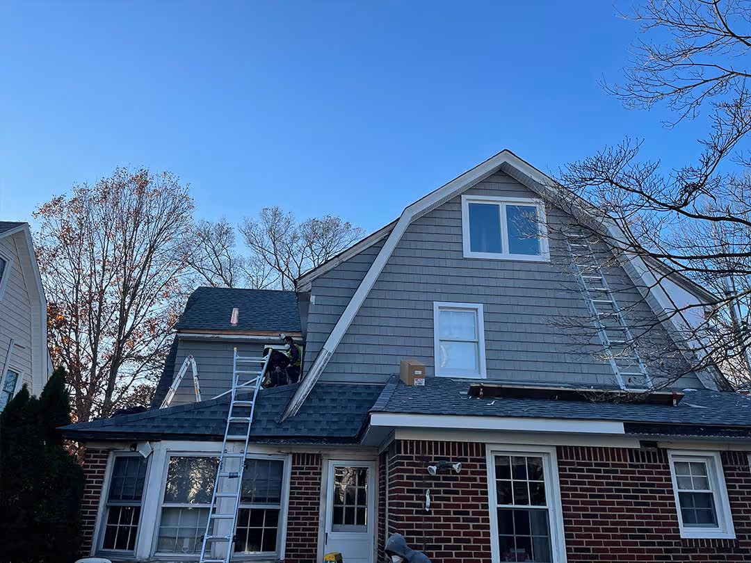 Two ladders leaning on a house with gray siding and brick lower walls under a clear blue sky.