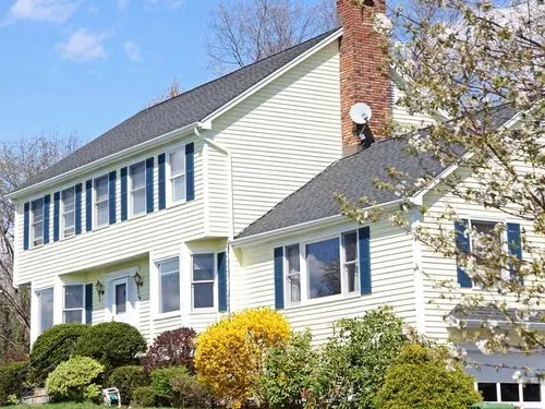 Two-story white house with vinyl siding, multiple windows, chimney, and green bushes in front yard.
