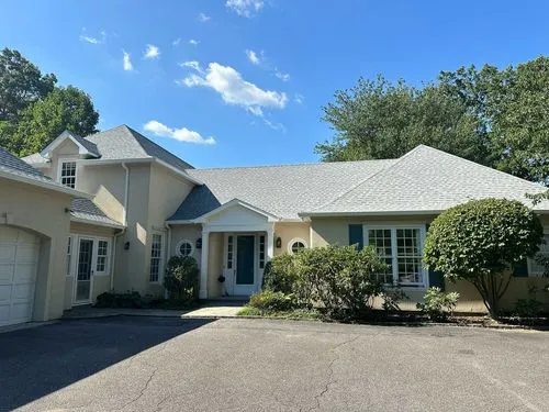 Single-story beige house with white trim, multiple windows, and surrounding green trees under a blue sky.