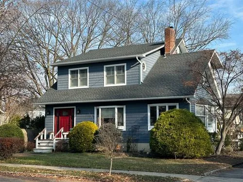 Two-story blue house with white trim, red front door, and surrounding bushes under a clear sky.