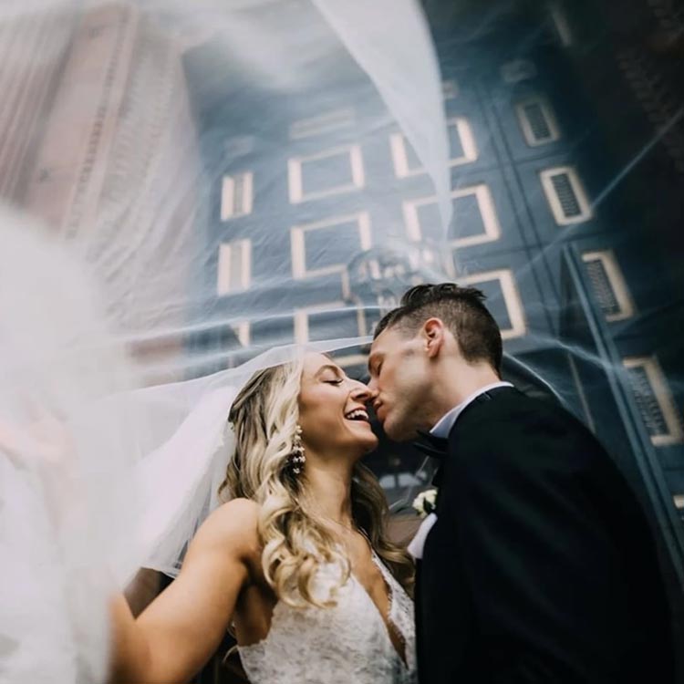 Bride and groom sharing a joyful kiss under veil with dramatic city architecture in the background