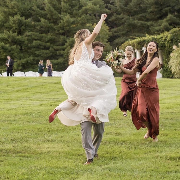 Joyful bride being carried by groom with bridesmaids celebrating in an open field wedding