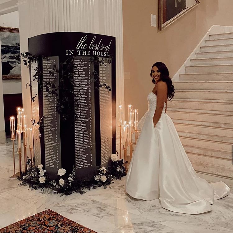 Bride in satin gown standing beside dramatic candlelit wedding seating chart with ‘the best seat in the house’ display