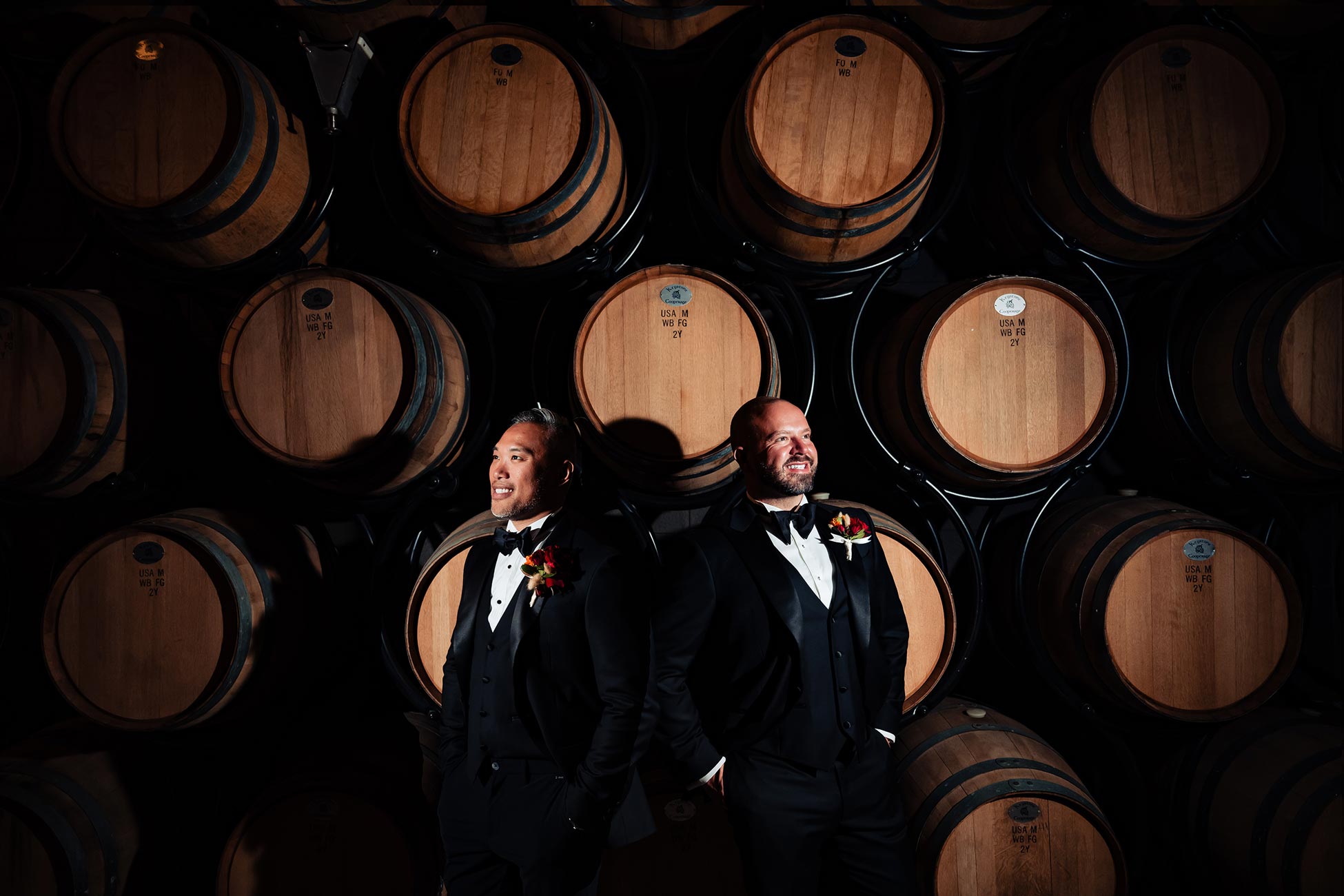 Two grooms in tuxedos posing in front of wine barrels at their winery wedding celebration