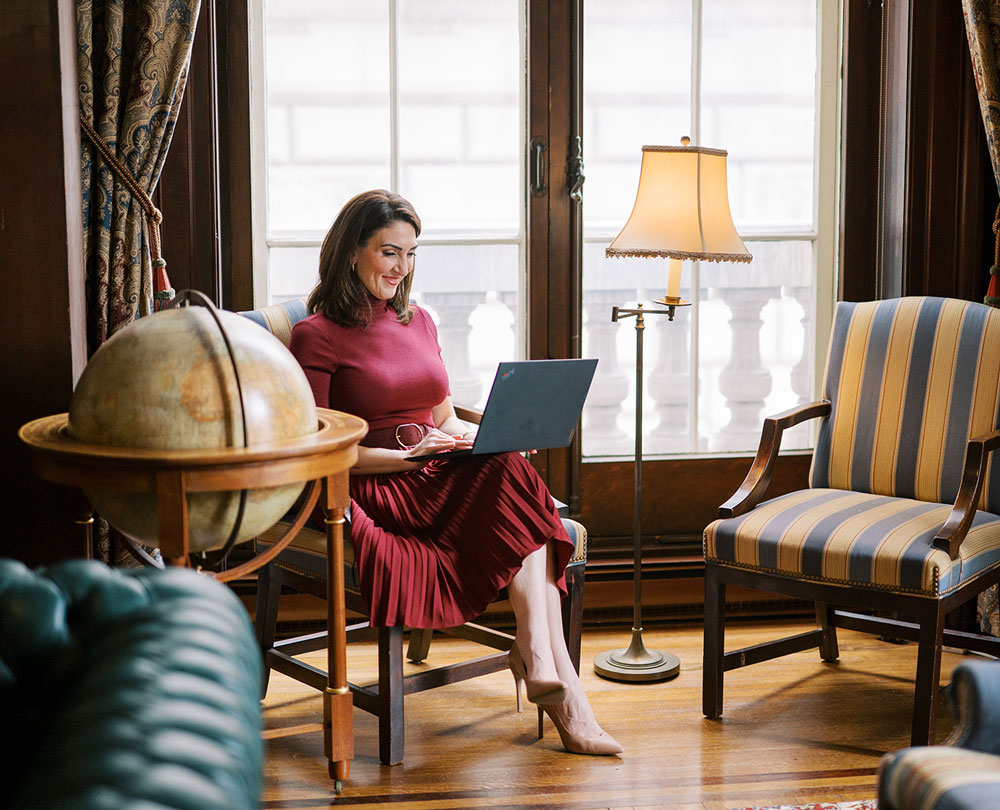 Marian Braccia working on her laptop in a historic library lounge wearing a maroon dress and heels