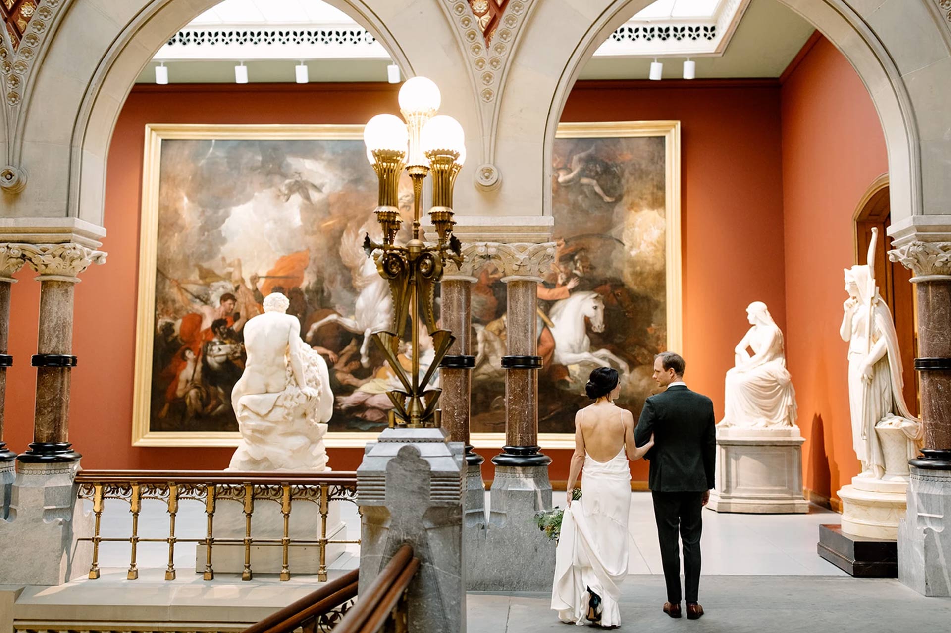 Bride and groom walking arm-in-arm through a grand art museum surrounded by sculptures and classical paintings.