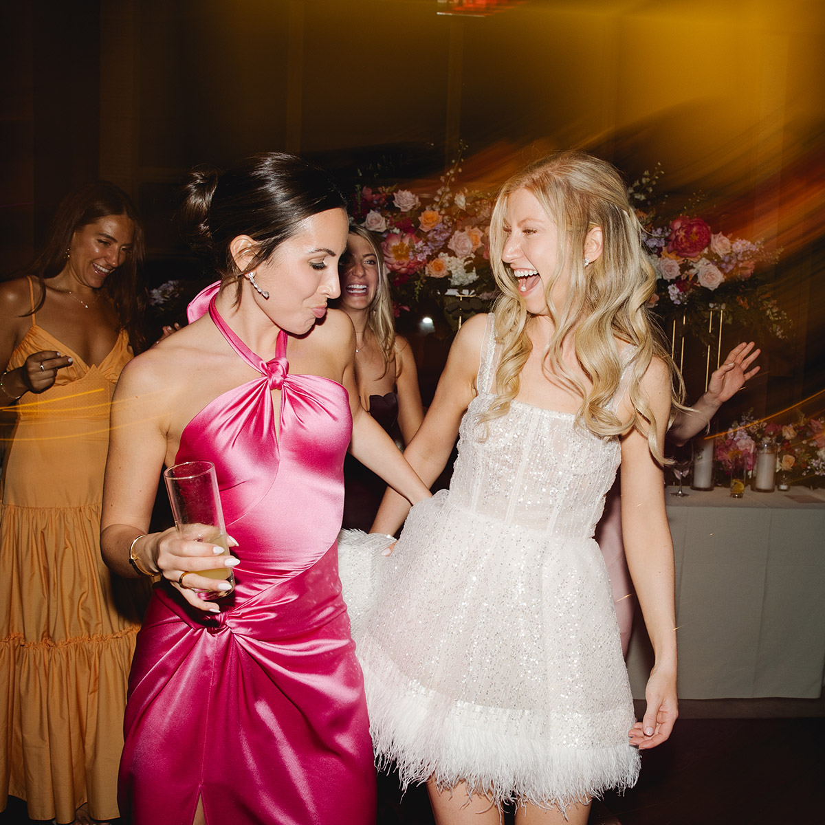 Bride and guest dancing joyfully at a wedding reception, surrounded by warm lights and vibrant florals.