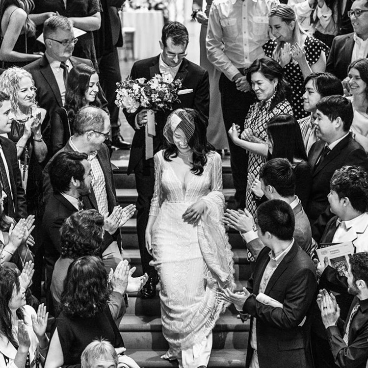 Bride in a vintage-inspired gown and veil walking down a staircase, surrounded by applauding guests in an elegant black and white moment.