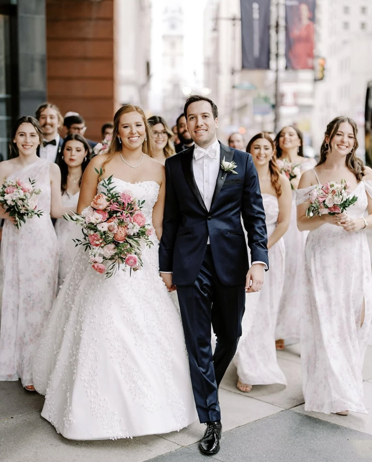 Bride and groom walking joyfully with their wedding party through a city street, surrounded by bridesmaids in soft floral dresses and groomsmen in classic tuxedos.