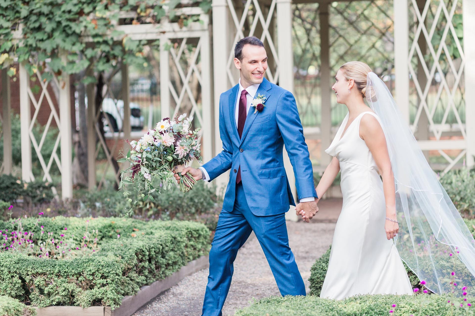 Bride and groom walking hand-in-hand through a garden, smiling joyfully as the groom carries a bouquet.