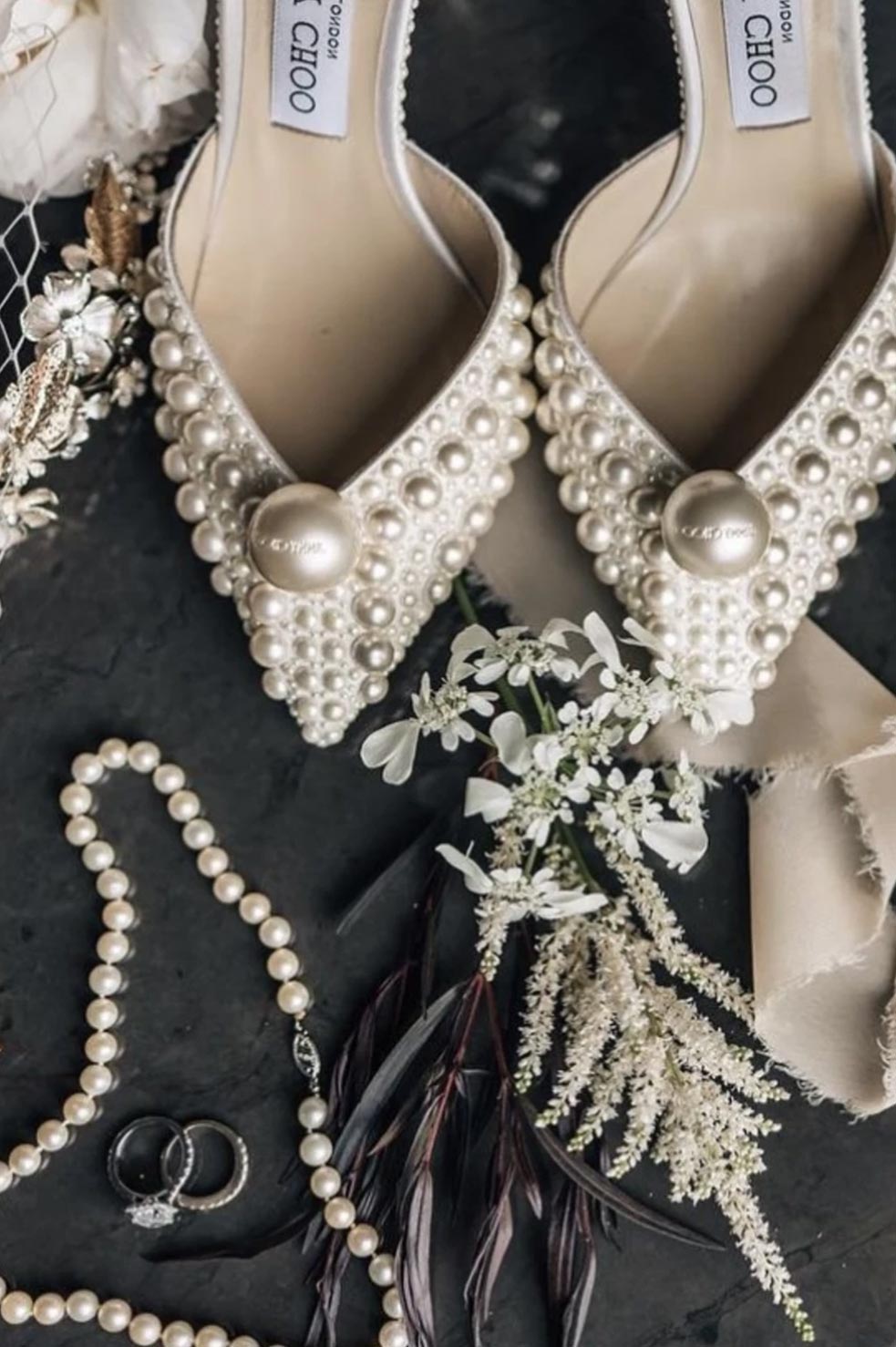 Flat lay of elegant bridal details: pearl-embellished Jimmy Choo heels, a pearl necklace, wedding rings, delicate white florals, and soft neutral ribbons — a timeless touch of sophistication.