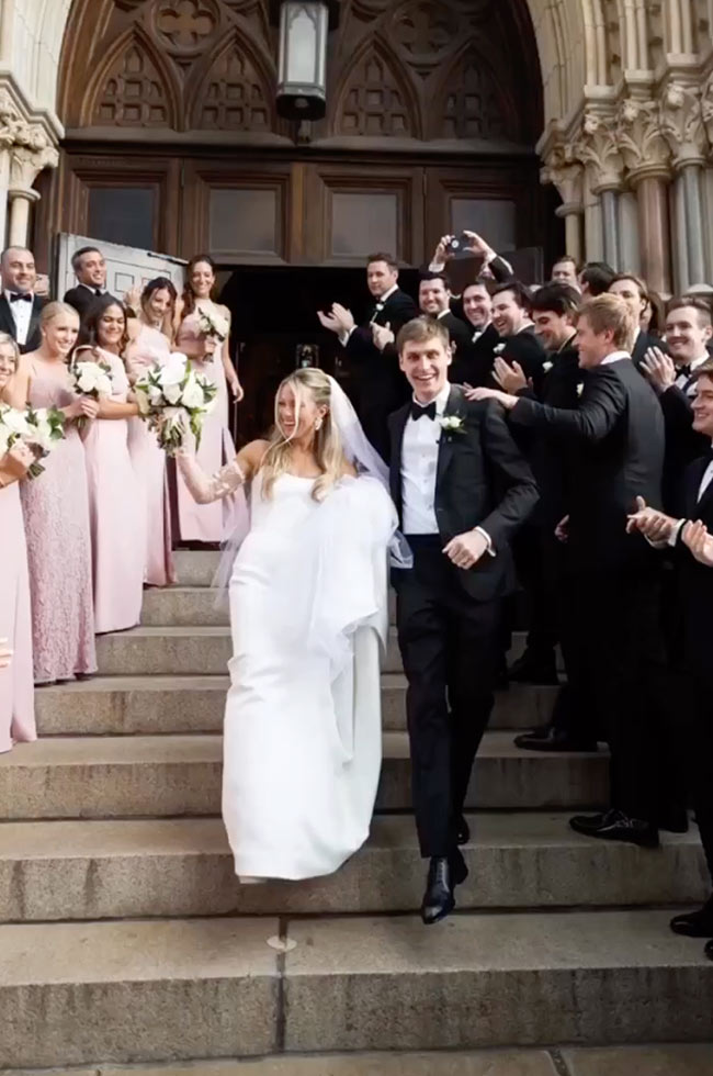 Bride and groom joyfully exit a grand cathedral surrounded by cheering bridesmaids in blush dresses and groomsmen in black tuxedos.