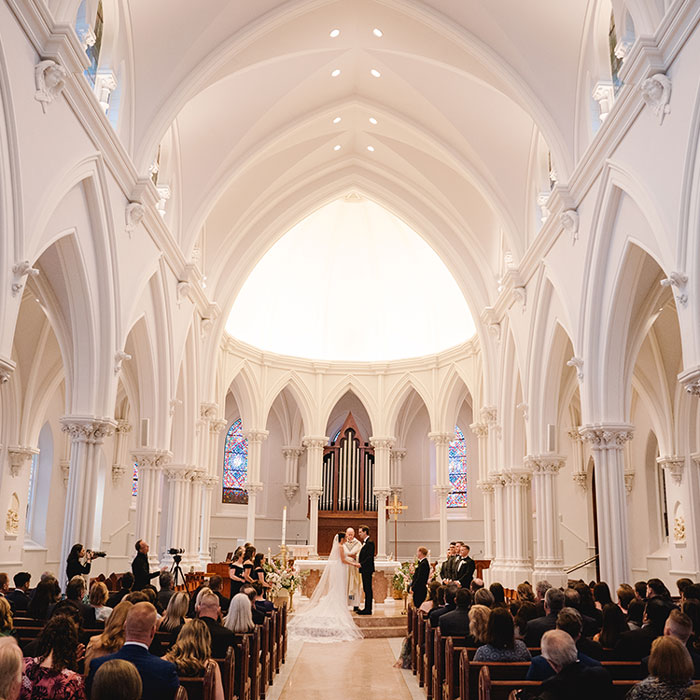 Elegant church wedding ceremony captured under soaring arches and soft golden light, with the couple centered at the altar before their gathered guests.