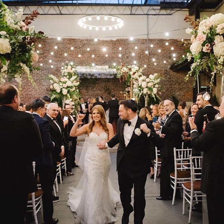 Joyful newlyweds walk back up the aisle under twinkling bistro lights, surrounded by cheering guests and towering floral arrangements