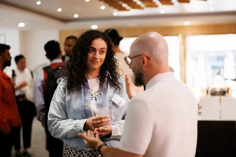A woman with curly hair holding a drink and listening attentively to a bald man wearing glasses at the reception for the Edtech Engine Demo Day.
