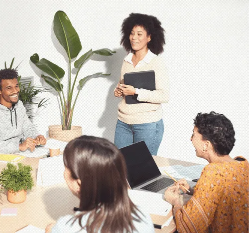 Four diverse coworkers engaged in a meeting with one standing woman holding a folder, smiling colleagues seated around a table with a laptop and plants.
