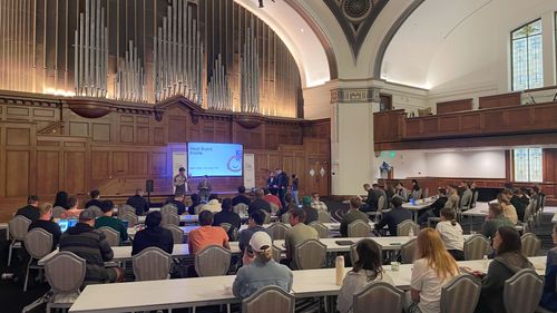 Large group of people seated and listening to four presenters speaking in a grand hall with wooden paneling and a large pipe organ in the background.