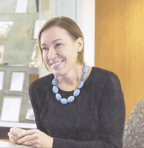 Smiling woman with short brown hair wearing a black top and a chunky light blue necklace seated indoors.