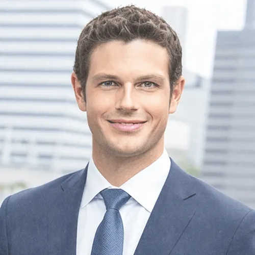 Portrait of a smiling man in a blue suit and tie with a blurred cityscape background.