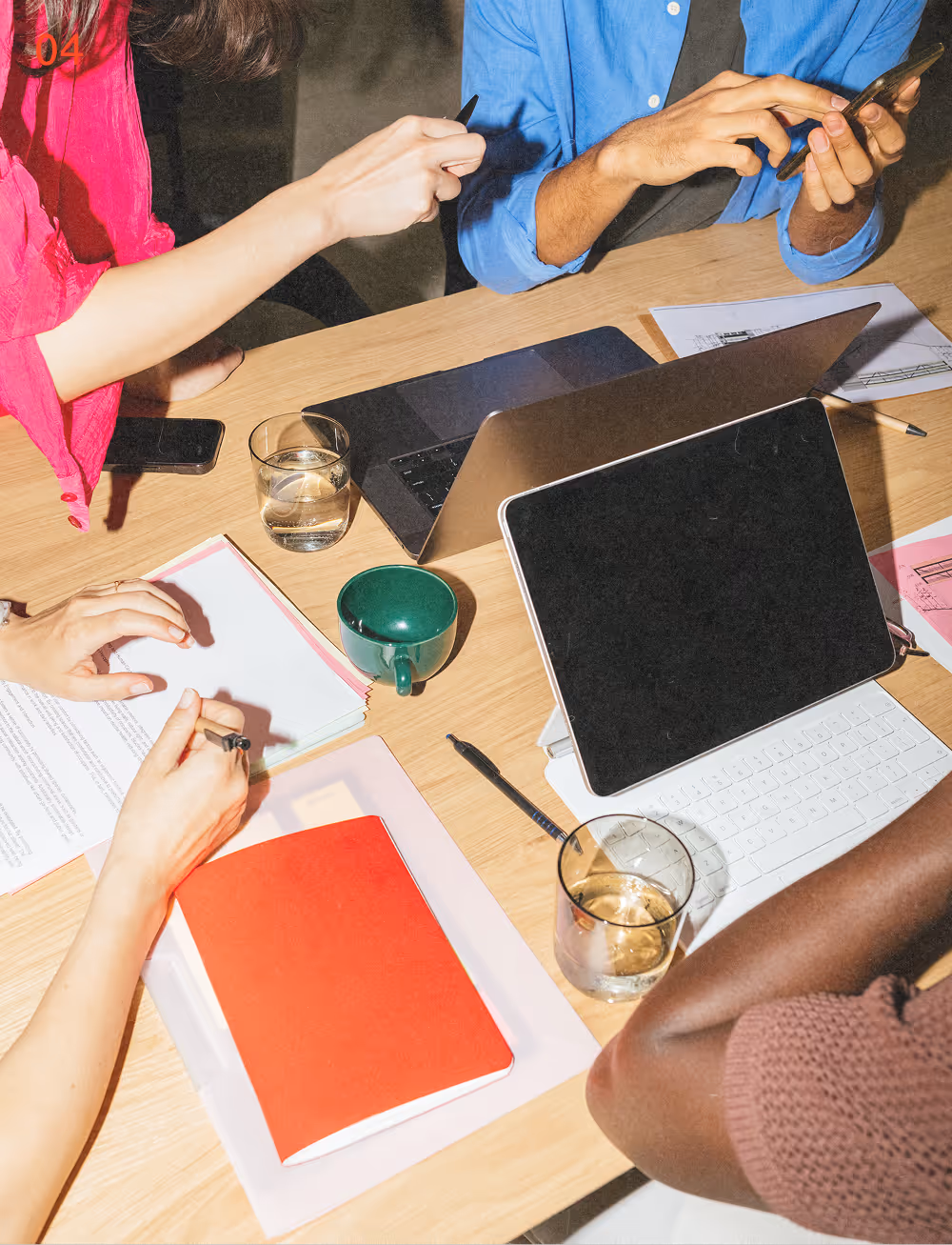 Four people collaborating at a wooden table with laptops, documents, a tablet, pens, and glasses of water.