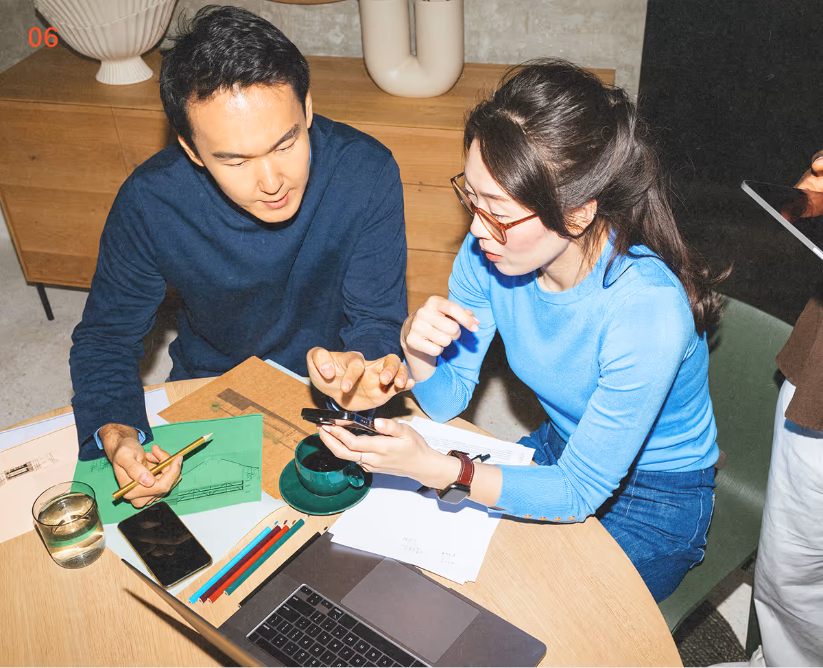 Two people collaborate at a table with colored pencils, a laptop, and a smartphone.