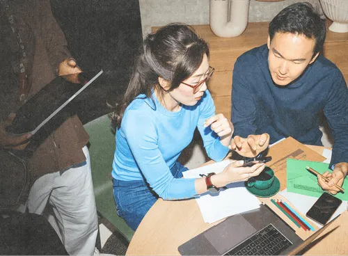Two people sitting at a table collaborating, one shows a phone screen while the other holds a pencil over documents and colored pencils, with a laptop and drinks on the table.