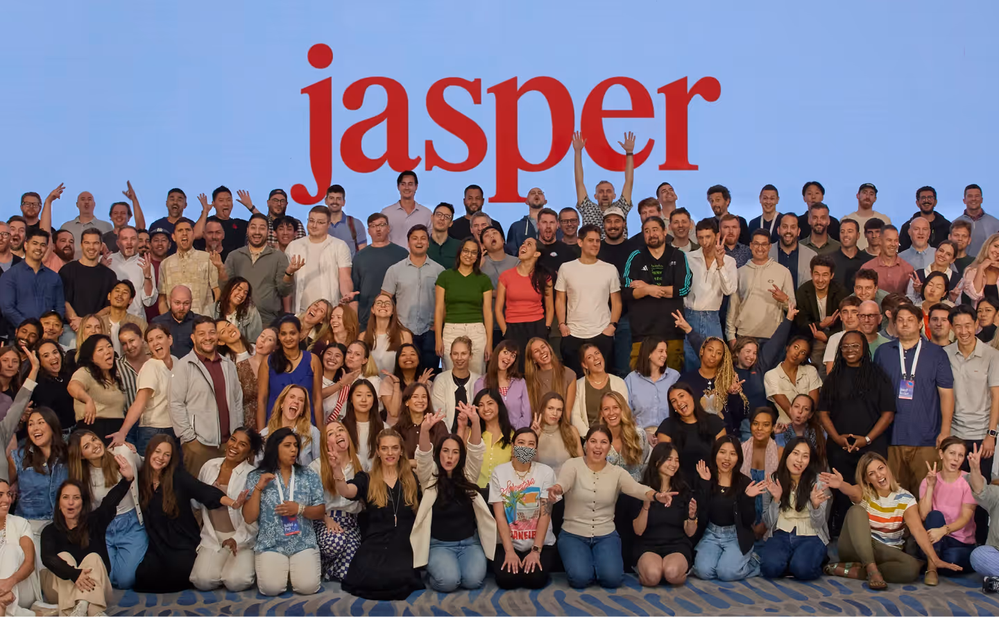 Large diverse group of people posing and smiling in front of a blue background with the red word ‘jasper’ above them.