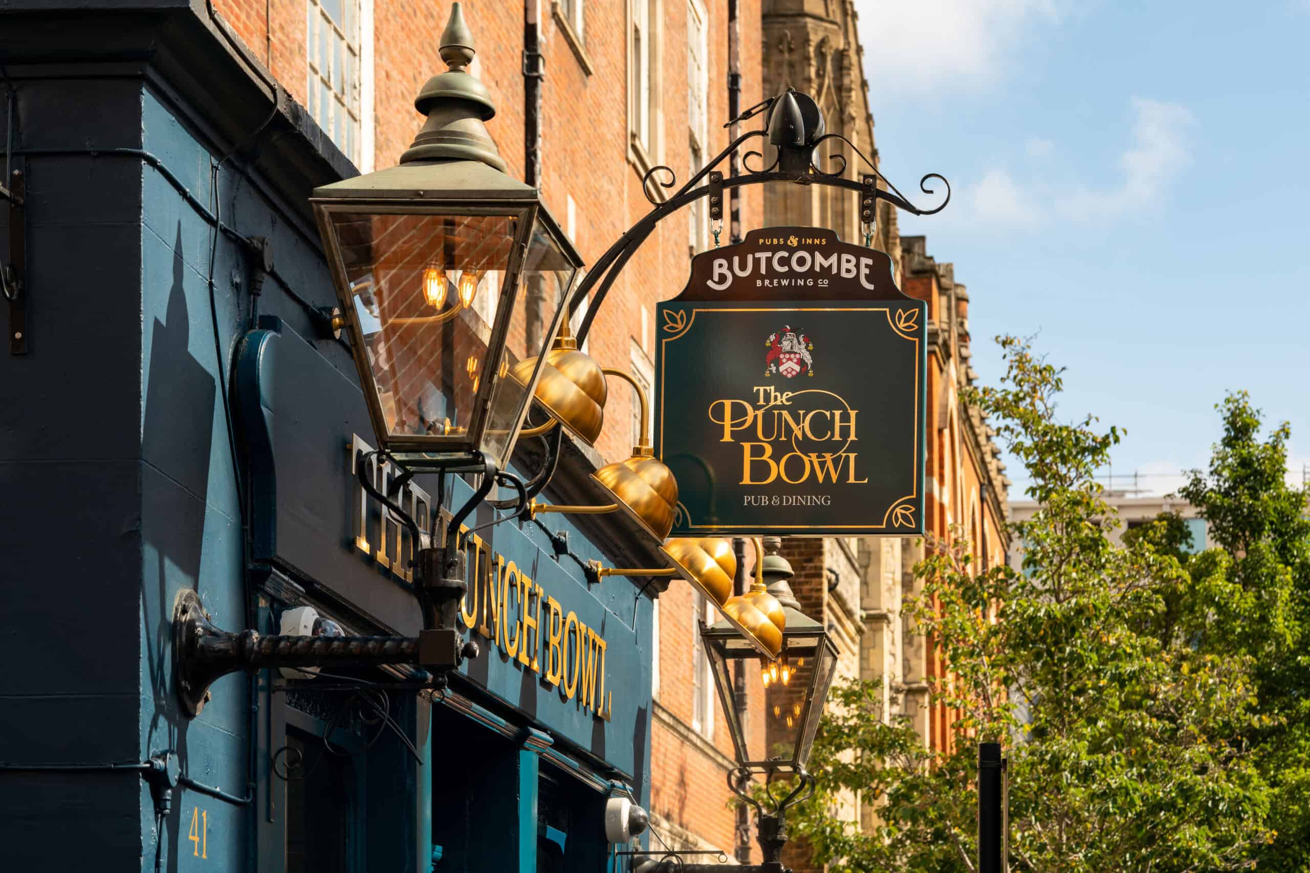 Street view of The Punch Bowl pub and dining sign hanging with vintage lamps on a brick building facade.