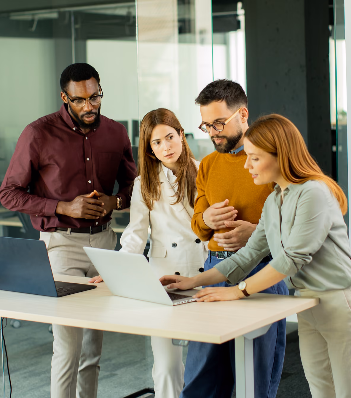 Four colleagues gather around a laptop at a standing desk as one points to the screen in a bright, modern office.
