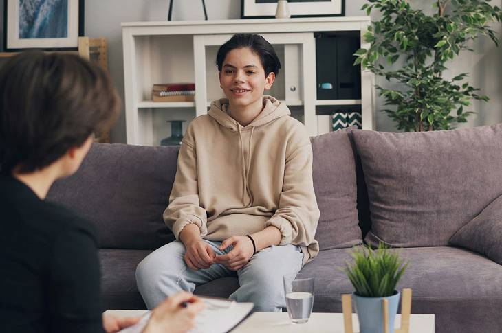 Teenager in a beige hoodie sitting on a couch and talking to a person holding a notepad in a cozy living room.
