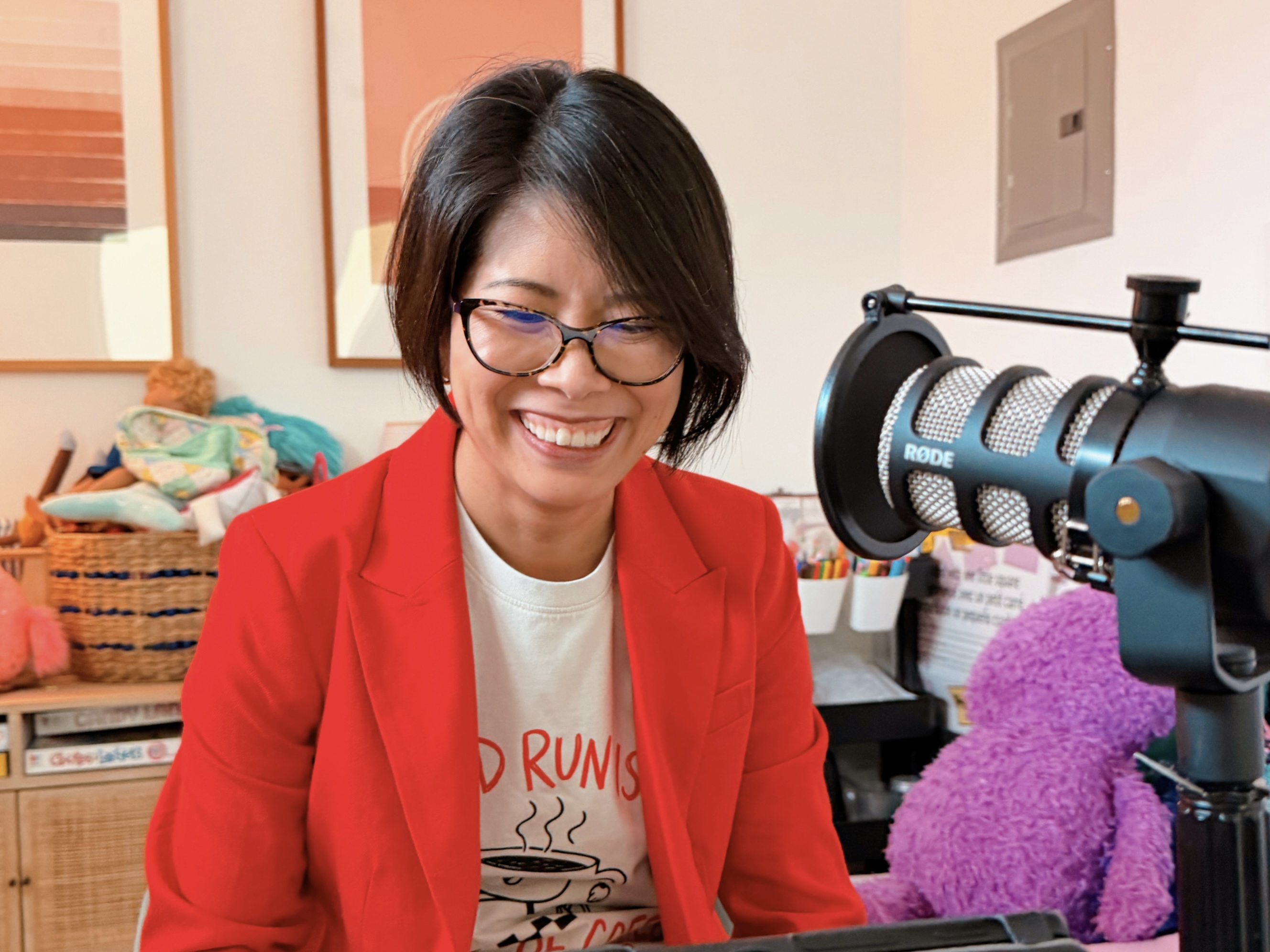 Smiling woman in glasses and red jacket recording with a microphone in a cozy room.