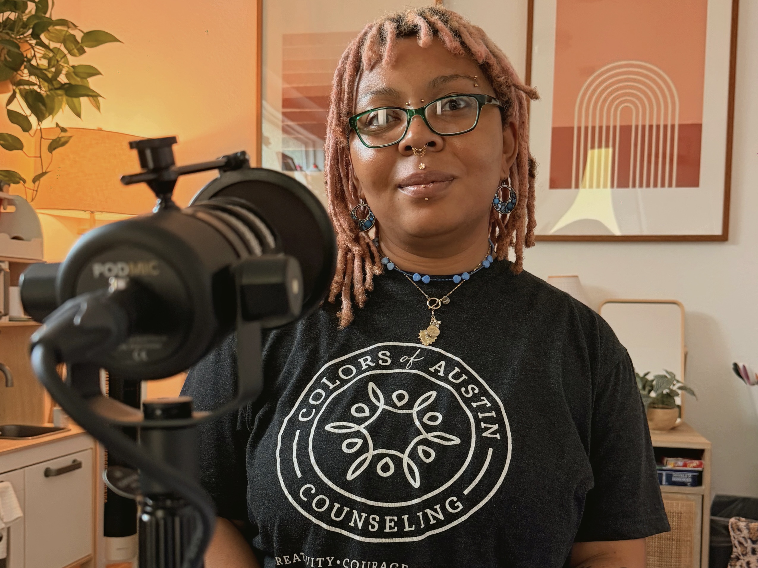 Person with pink dreadlocks and glasses speaking into a microphone wearing Colors of Austin Counseling shirt.
