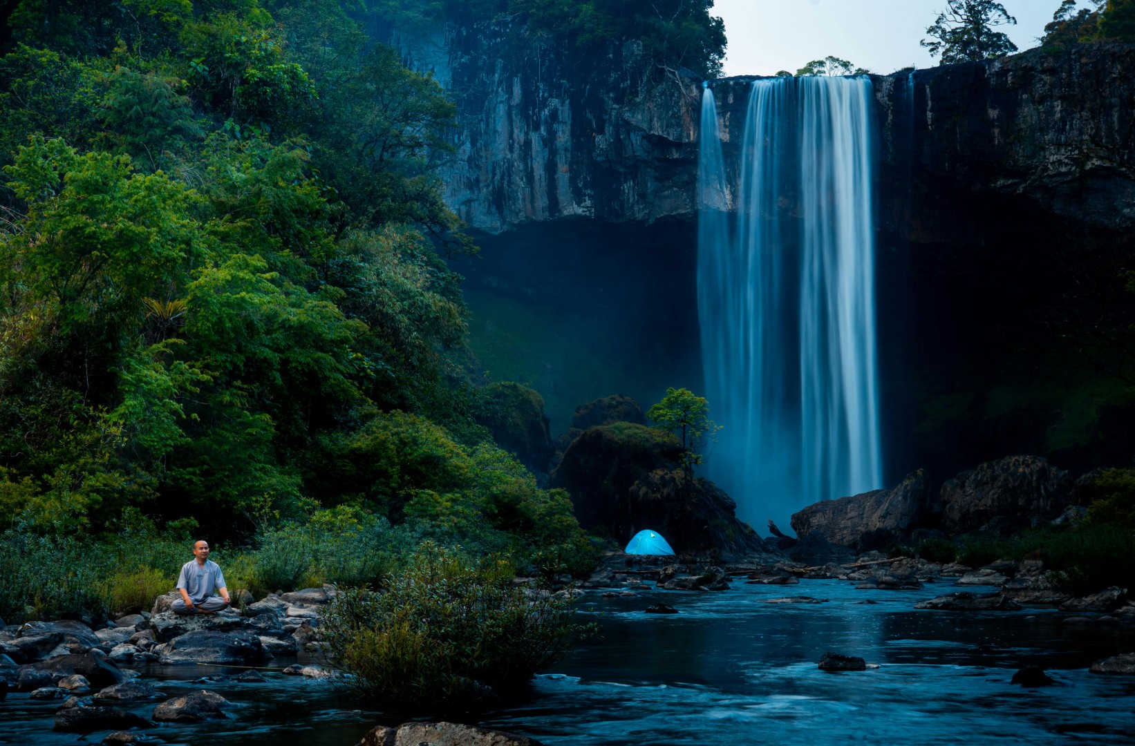 Person meditating on rocks near a flowing waterfall surrounded by lush green trees and a blue tent in the background.