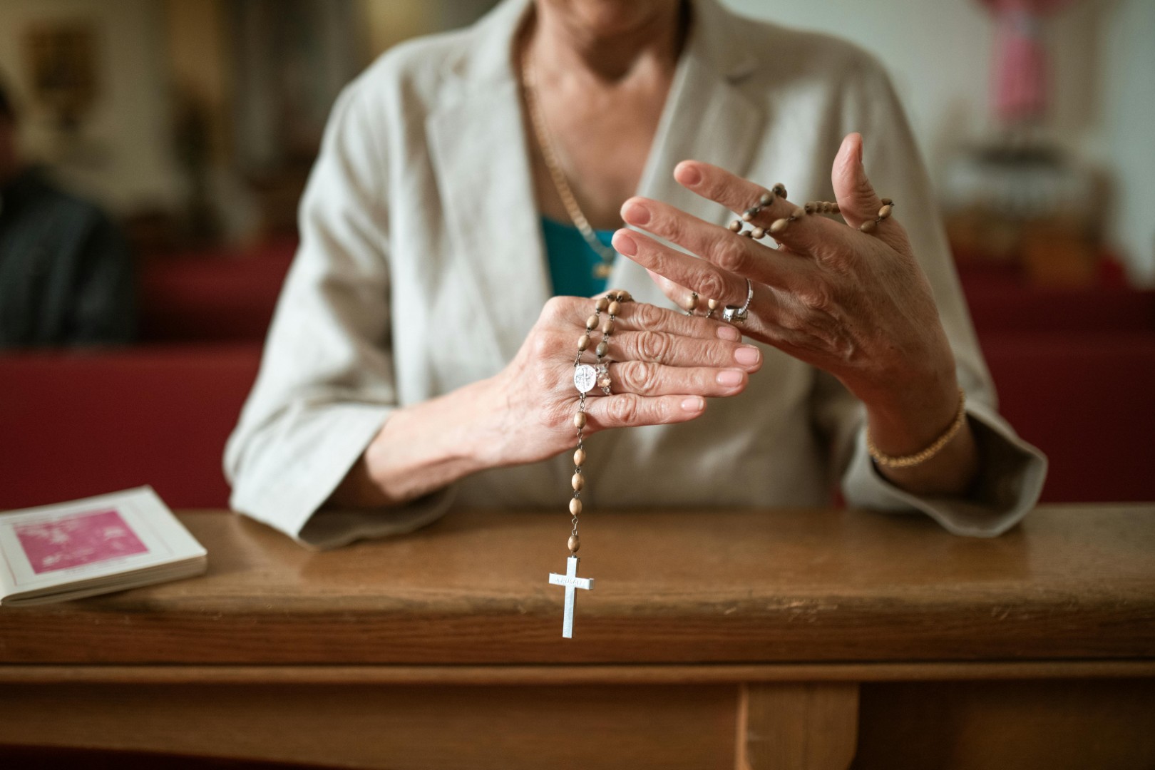 Person holding wooden rosary beads with a silver cross, sitting at a wooden pew with a prayer book nearby.
