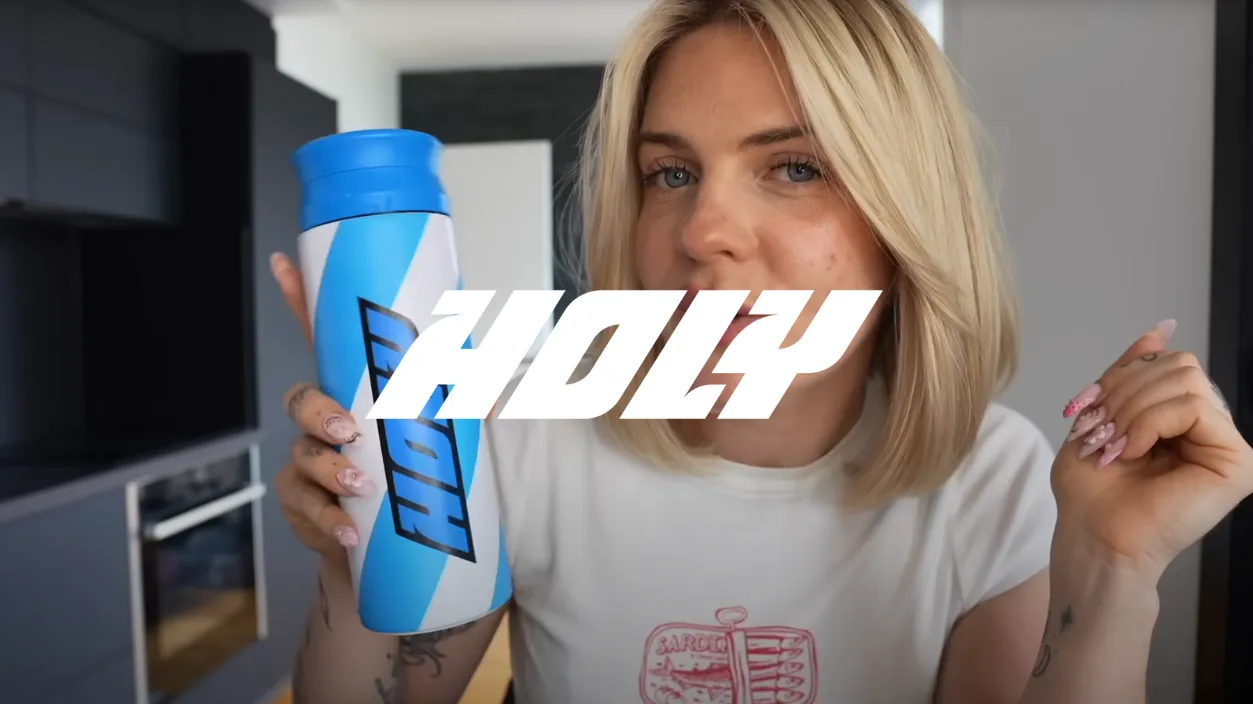 Jeune femme blonde tenant une bouteille bleue et blanche avec le logo HOLY dans une cuisine moderne.