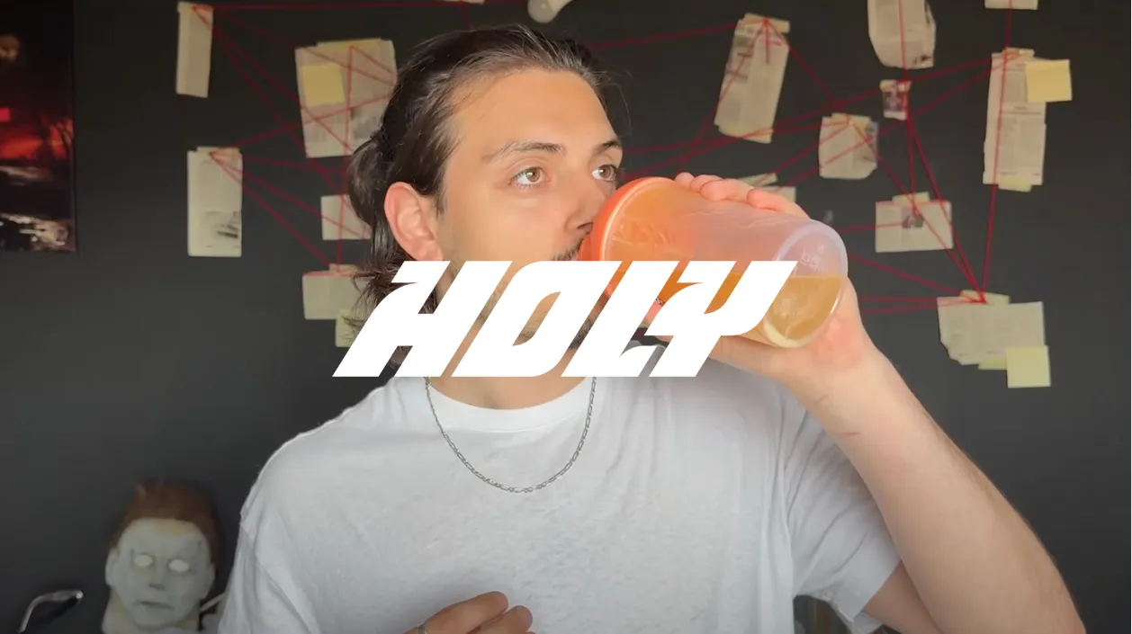 Un homme en t-shirt blanc boit d'un gobelet orange avec un mur noir en arrière-plan couvert de papiers reliés par des fils rouges.