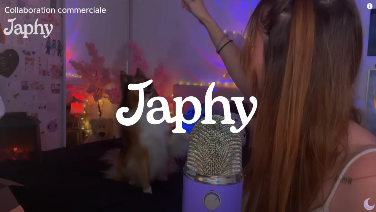 Une femme aux cheveux longs regarde un petit chien sur un lit dans une pièce décorée avec des lumières tamisées et un microphone violet au premier plan.