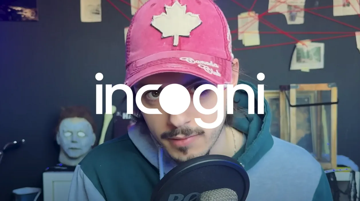 Un homme portant une casquette rose avec une feuille d’érable blanche et un sweat à capuche, parlant devant un microphone, avec un masque effrayant en arrière-plan.