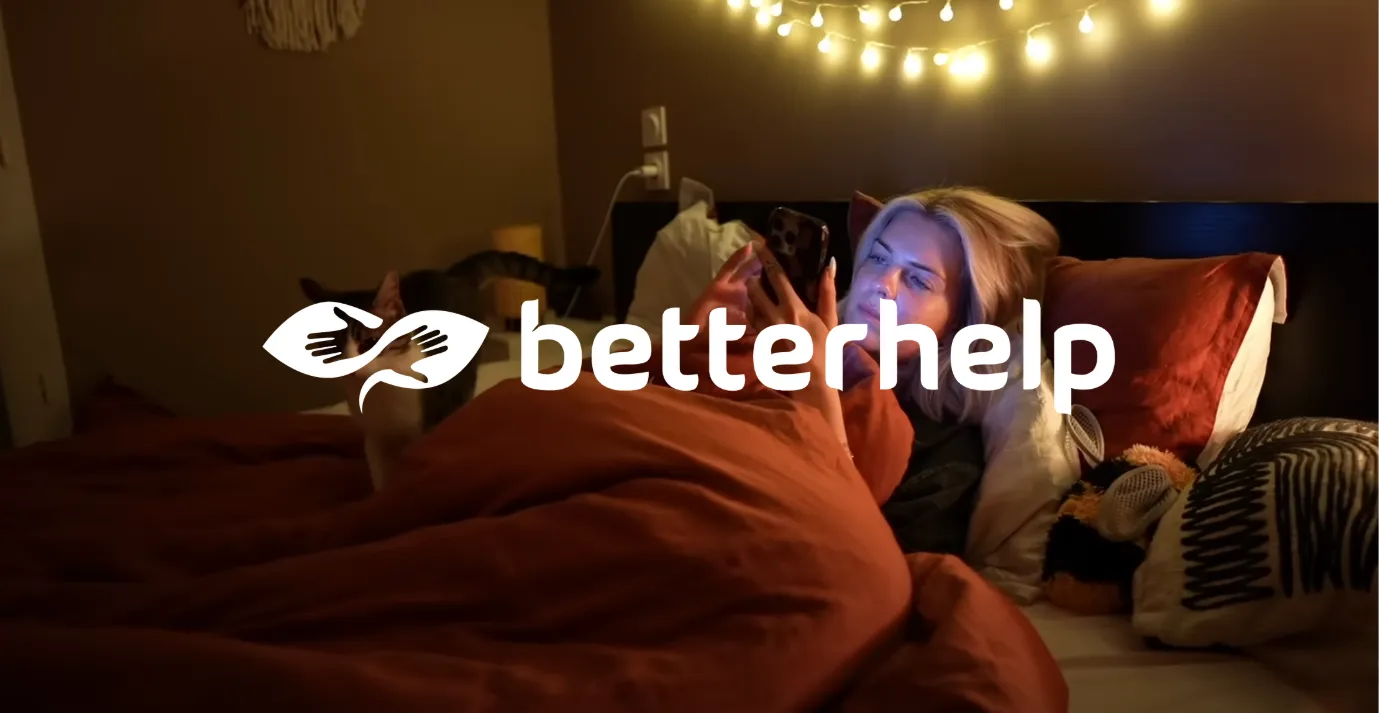 Personne allongée dans un lit avec une couverture orange, regardant un smartphone sous des guirlandes lumineuses suspendues.