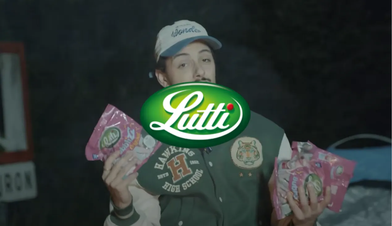 Un homme portant une casquette et une veste de style universitaire tenant plusieurs paquets de bonbons Lutti roses, sur un fond sombre.