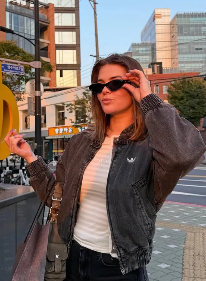 Femme portant une veste noire, un t-shirt blanc et des lunettes de soleil noires, posant dans une rue urbaine au coucher du soleil.