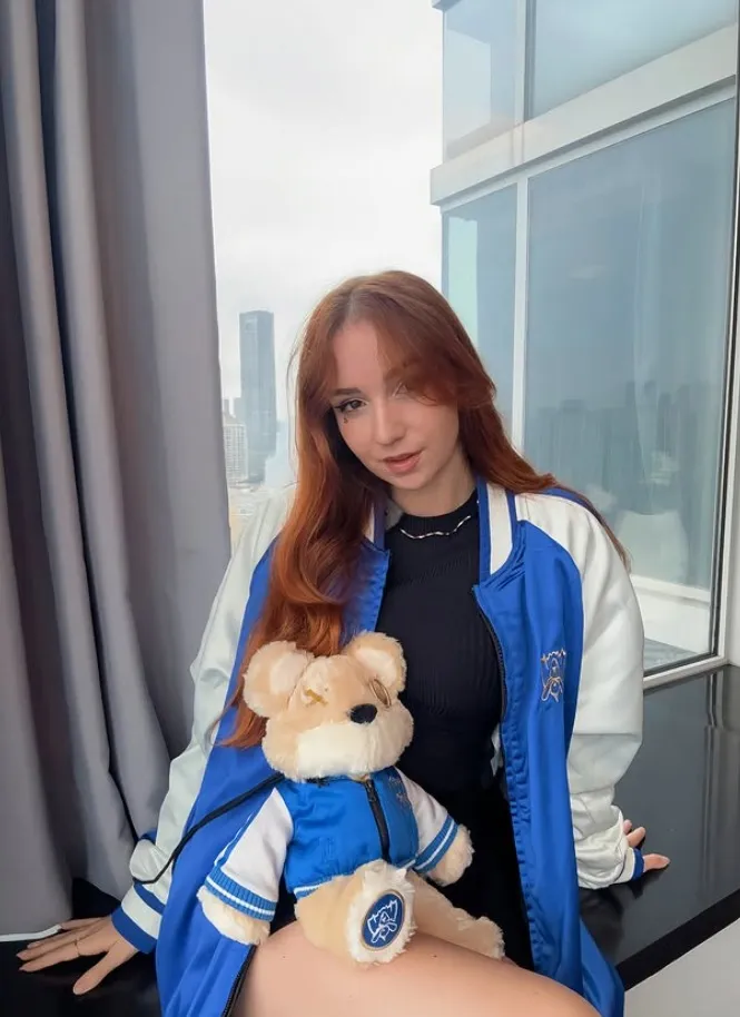 Jeune femme aux cheveux roux portant une veste bleu et blanc, assise avec un ours en peluche vêtu d'une veste assortie sur ses genoux, devant une fenêtre avec une vue urbaine.