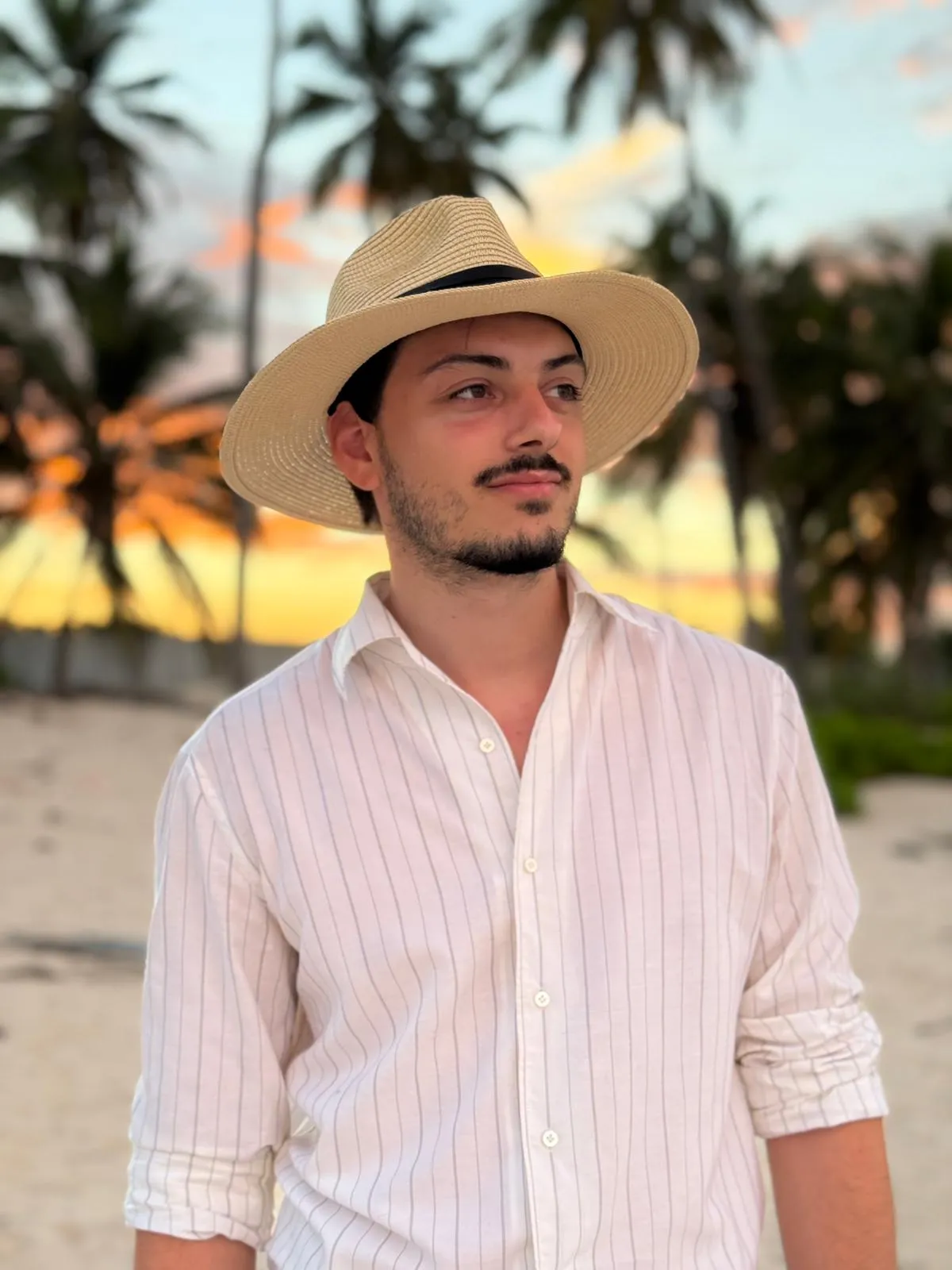 Jeune homme portant un chapeau de paille et une chemise blanche à rayures, posant sur une plage avec des palmiers au coucher du soleil.