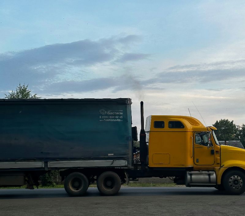 Yellow semi truck with covered flatbed trailer parked roadside at dusk, silhouetted against cloudy sky and distant trees.