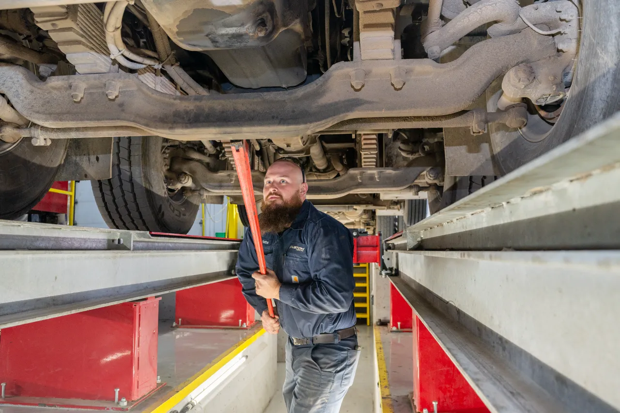 Diesel truck breakdown prevention inspection under lifted chassis with technician using pry bar in heavy-duty repair bay.