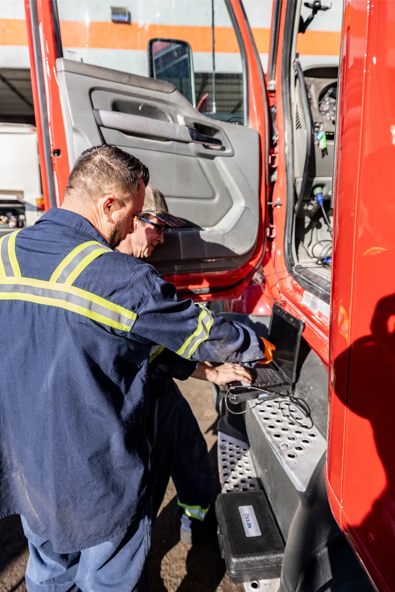 Technicians perform a pre trip inspection on a red heavy-duty truck using a laptop beside the open cab.