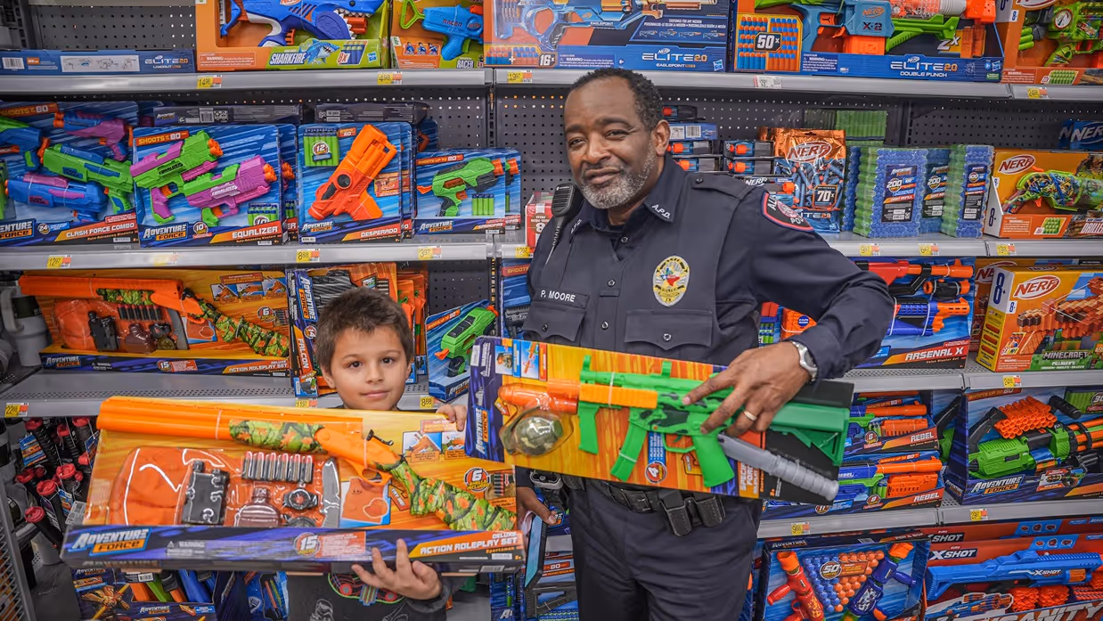 A police officer and a child hold colorful toy guns, surrounded by shelves filled with various toy weapons and accessories.